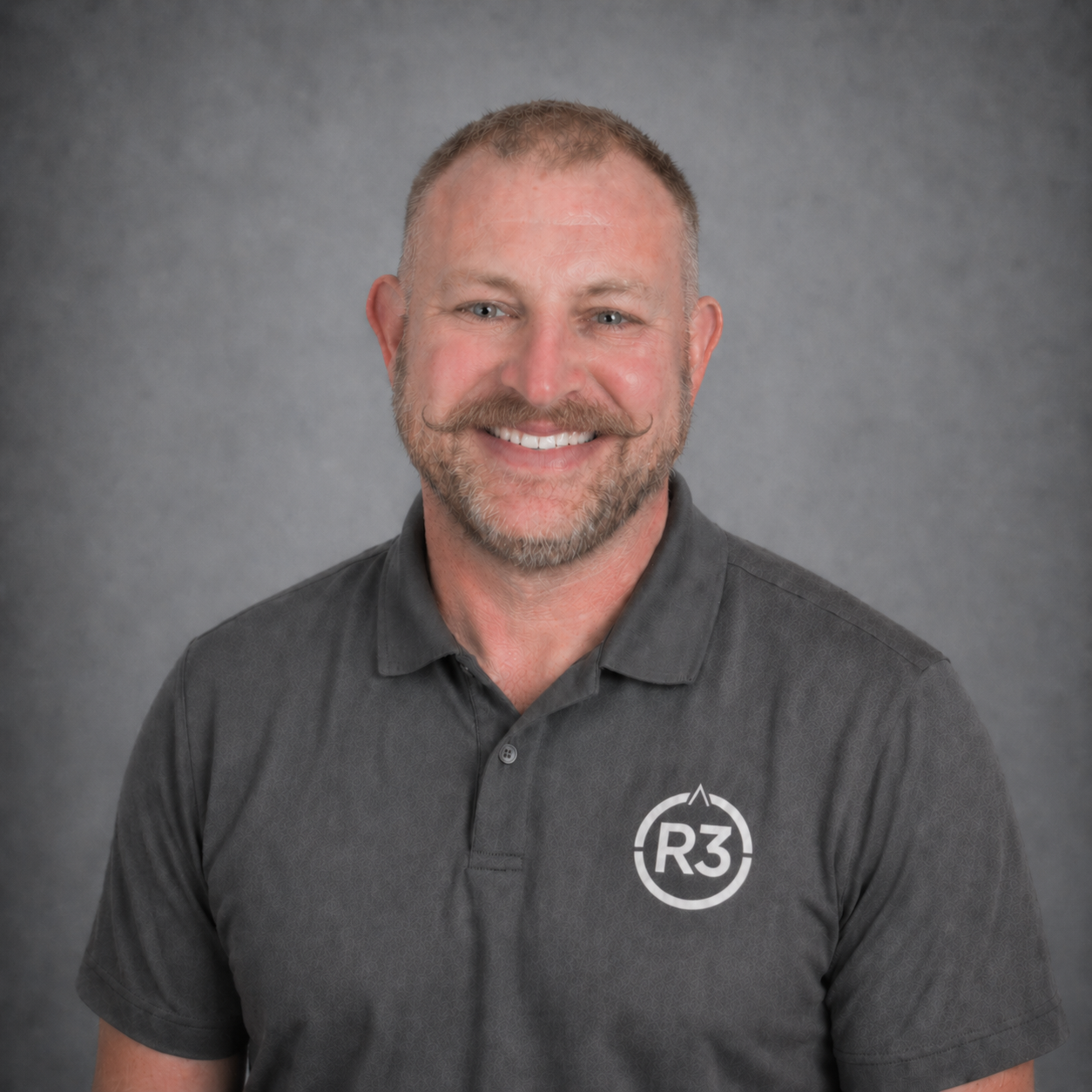 Bryan Syrett, professional head-and-shoulders portrait in a dark grey polo with the R3 logo, smiling at the camera against a neutral grey studio background.
