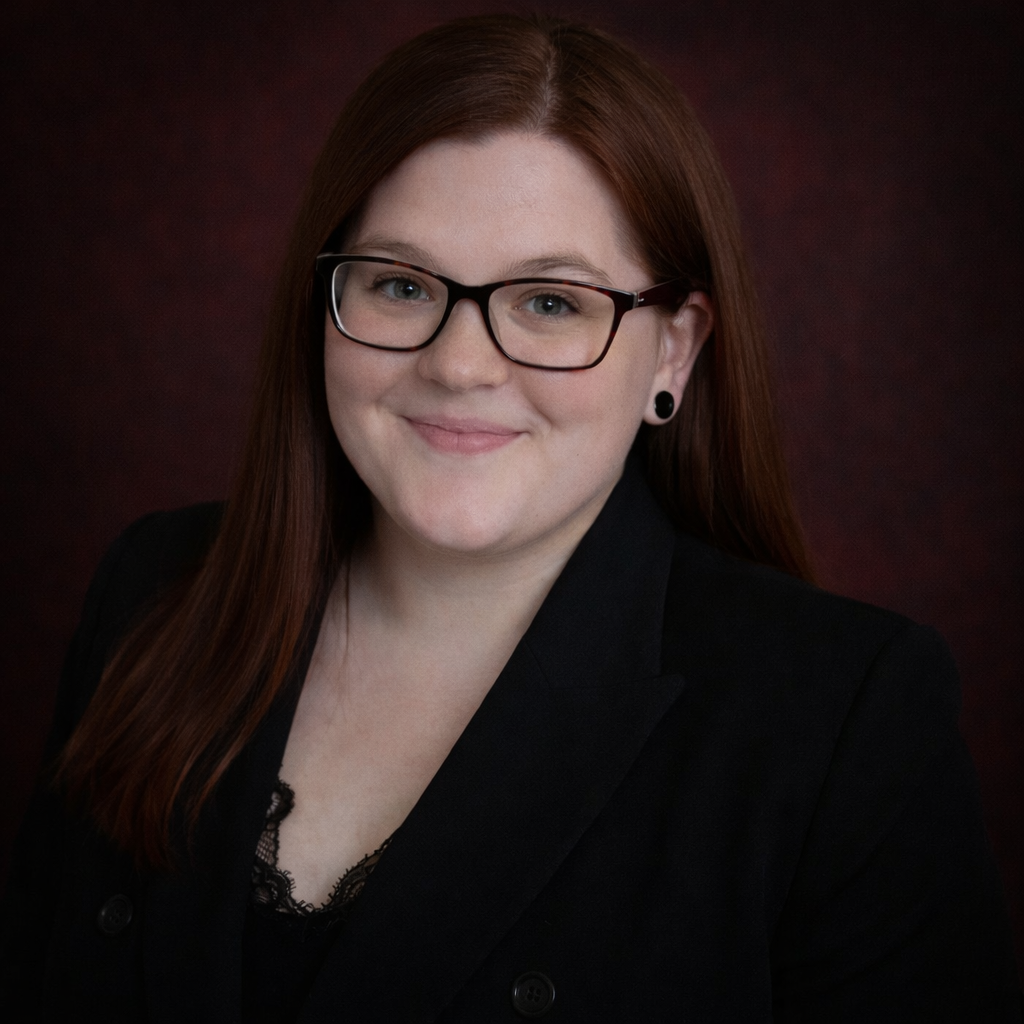 Lindsey Burnett, professional head-and-shoulders portrait in a dark grey polo with the R3 logo and glasses, smiling at the camera against a neutral grey studio background.